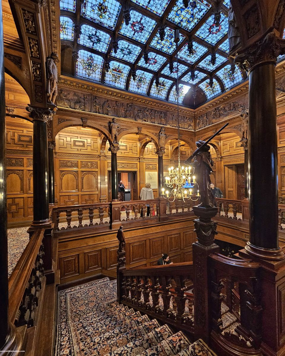 Carved wooden gallery of arches and columns under a stained-glass ceiling, patterned carpet running up the stairs