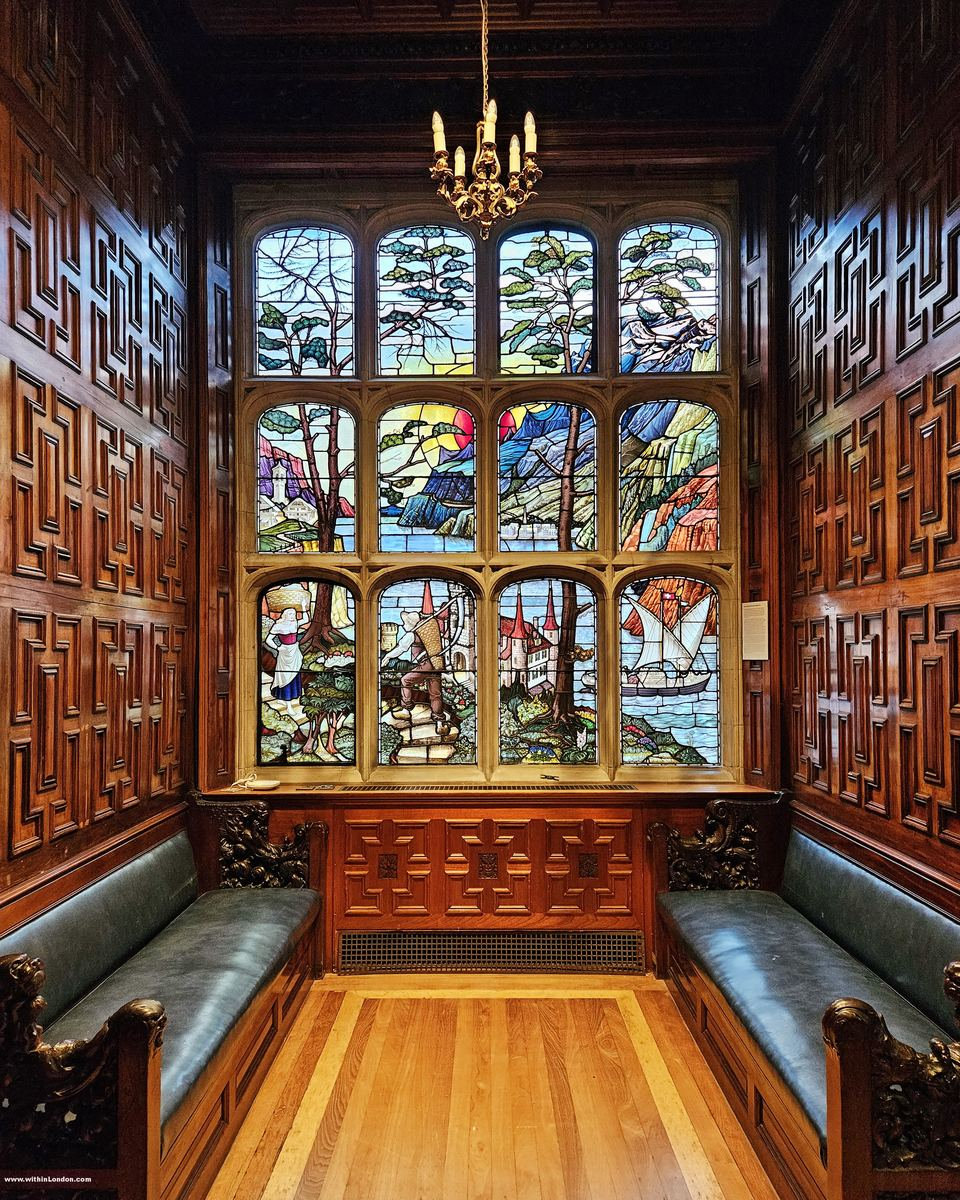 Window bay filled with colourful stained glass above dark carved panelling, green leather benches and a chandelier