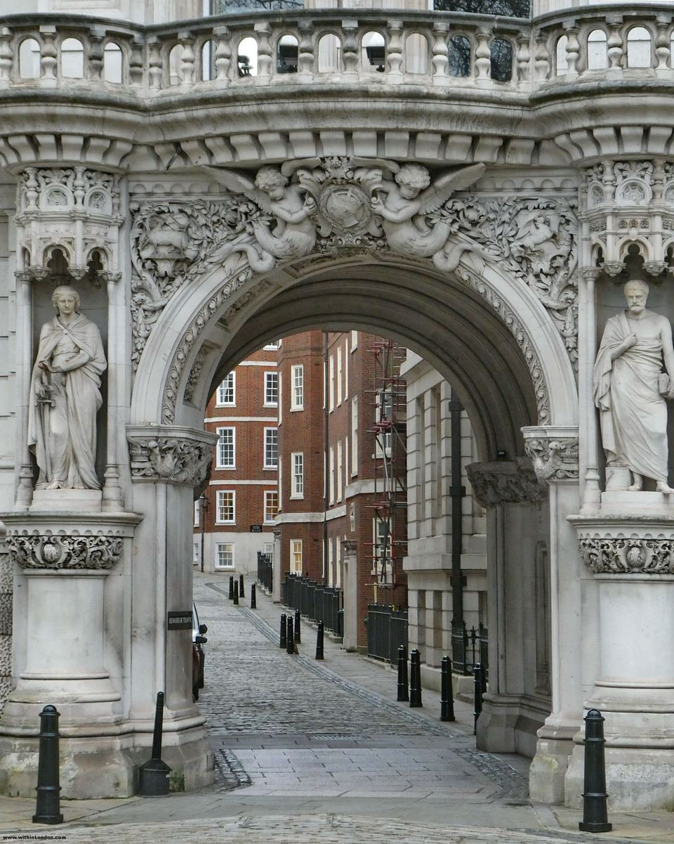 Ornate white stone archway with carved statues in niches on either side, a cobbled lane and red-brick buildings seen through it