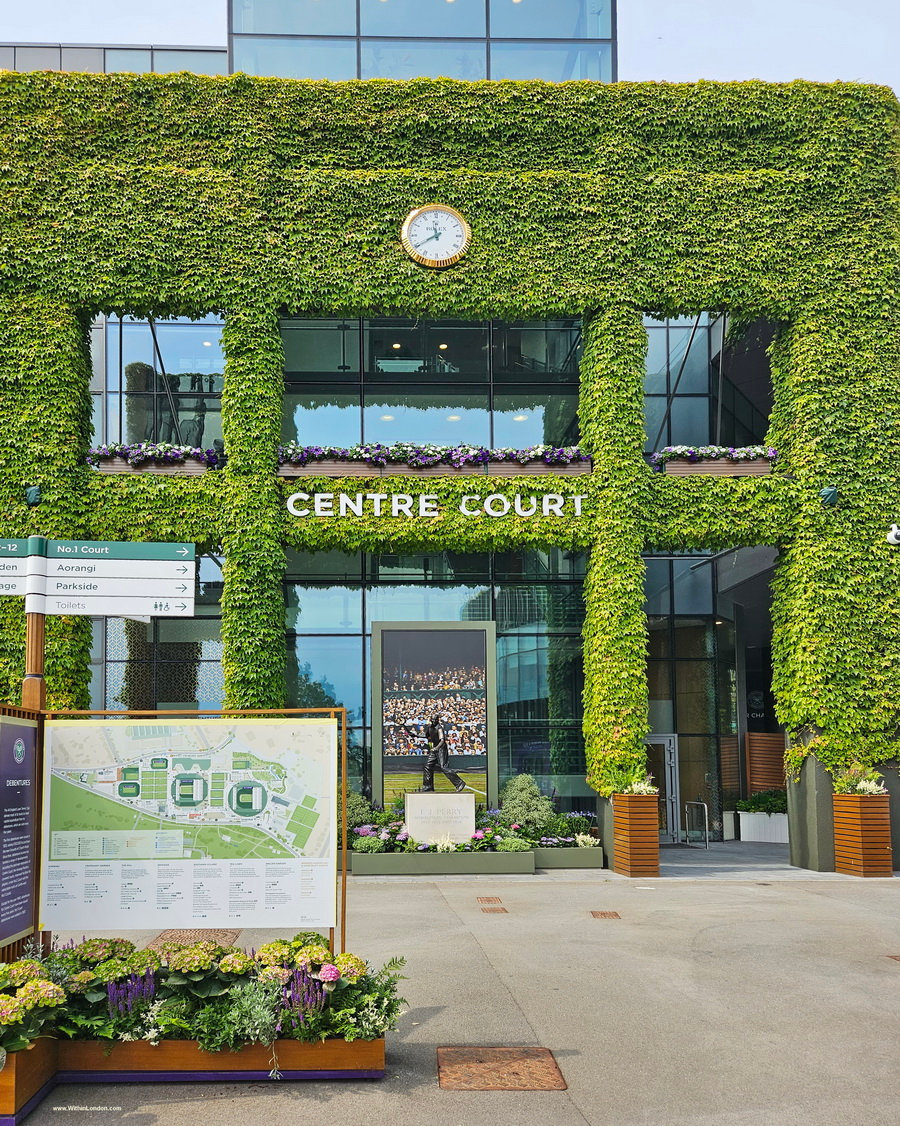 Ivy-covered facade of Centre Court with its clock and lettering, a grounds map sign and flower beds in front