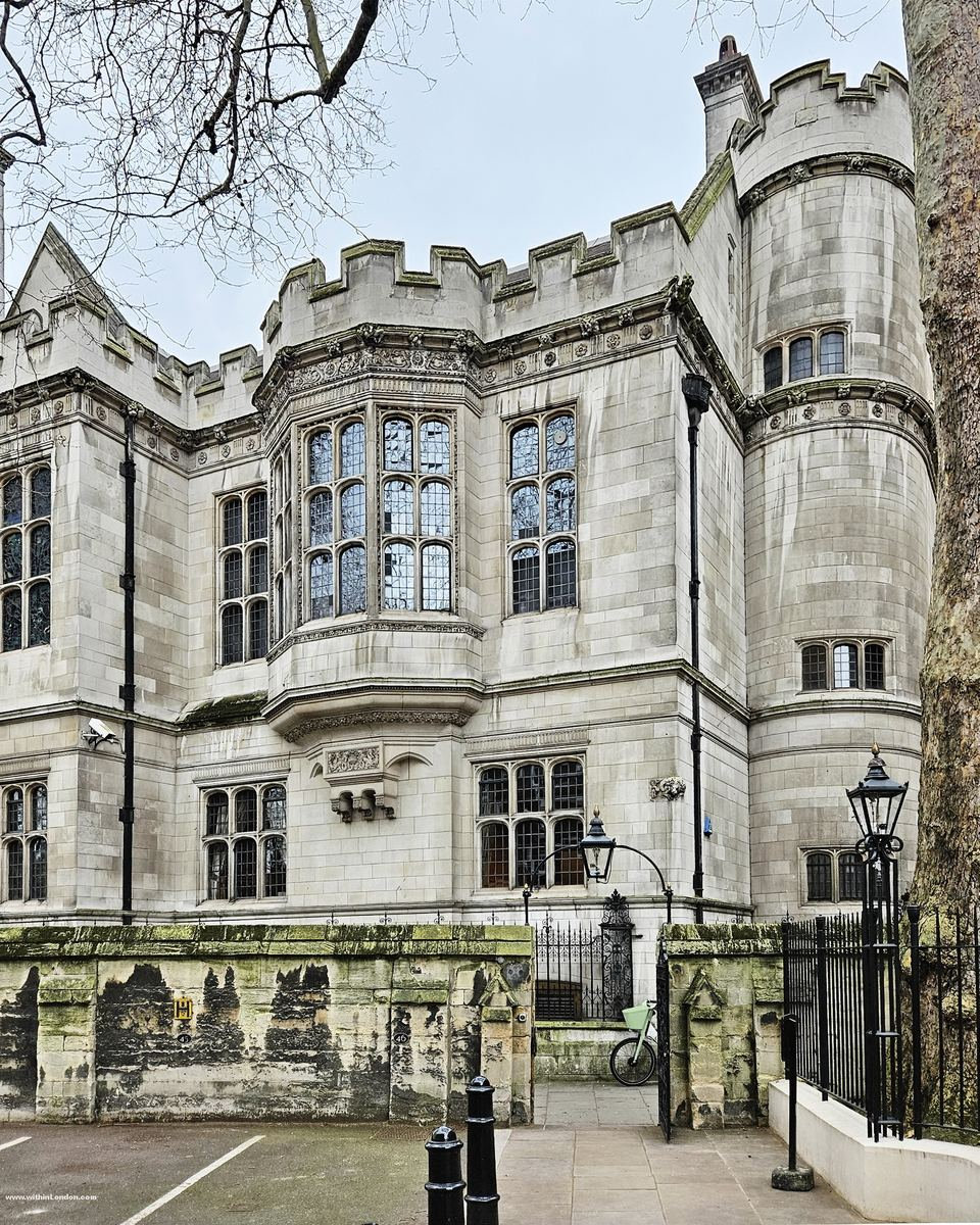 Pale stone castellated building with a bay window, black lamp posts and railings along the pavement