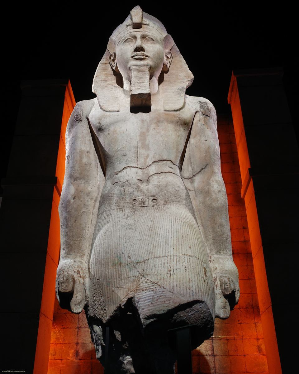Colossal limestone statue of Ramses II seated, bare-chested with striped nemes headdress and broad collar, lit from below against a dark background with orange architectural elements at Battersea Power Station
