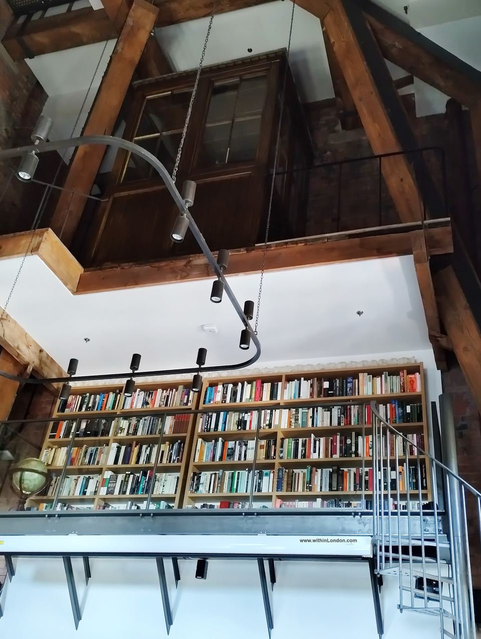 Tall white room with exposed timber beams, a wooden clock-winding box hanging high up, a gallery of bookshelves and a spiral staircase