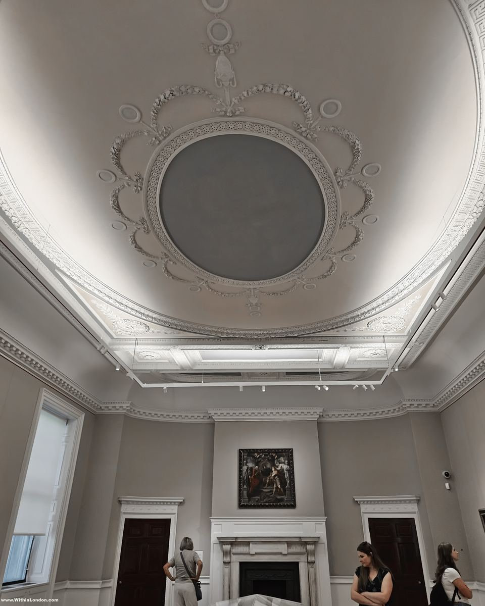 Tall white gallery with an ornate plaster ceiling framing a large oval, a single painting on the far wall and visitors standing below