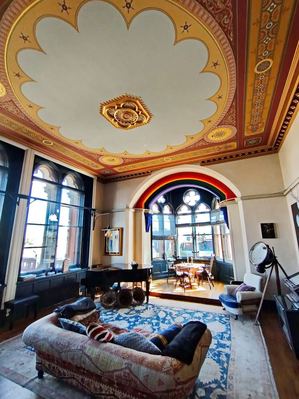 Drawing room with a scalloped cream ceiling framed in red and gold painted borders, a rainbow arch over the bay window and a grand piano