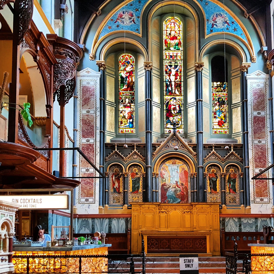 The apse at Mercato Mayfair: stained glass lancets and painted Romanesque arcading above a carved wooden reredos, with a bar counter and a gin cocktails sign in front of it
