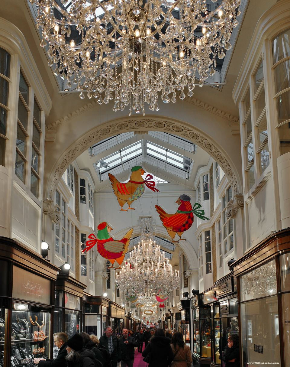 Burlington Arcade under its glass roof, with painted red and green birds hanging from the ceiling above shoppers and a long chandelier in the distance