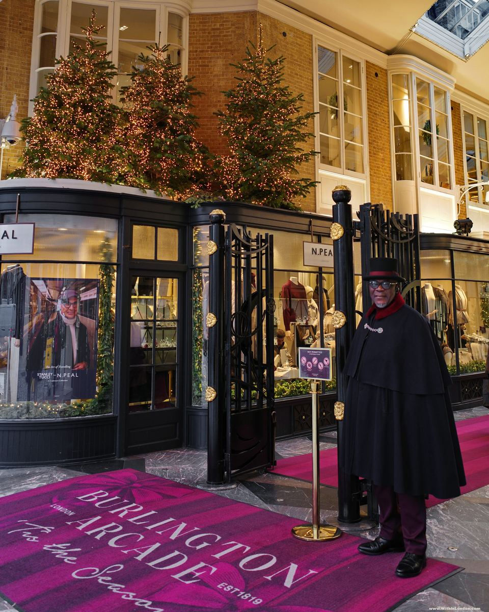 A beadle in a black cape and top hat standing beside a brass post and black iron gates, on a pink carpet printed Burlington Arcade, with lit fir trees above the shopfront