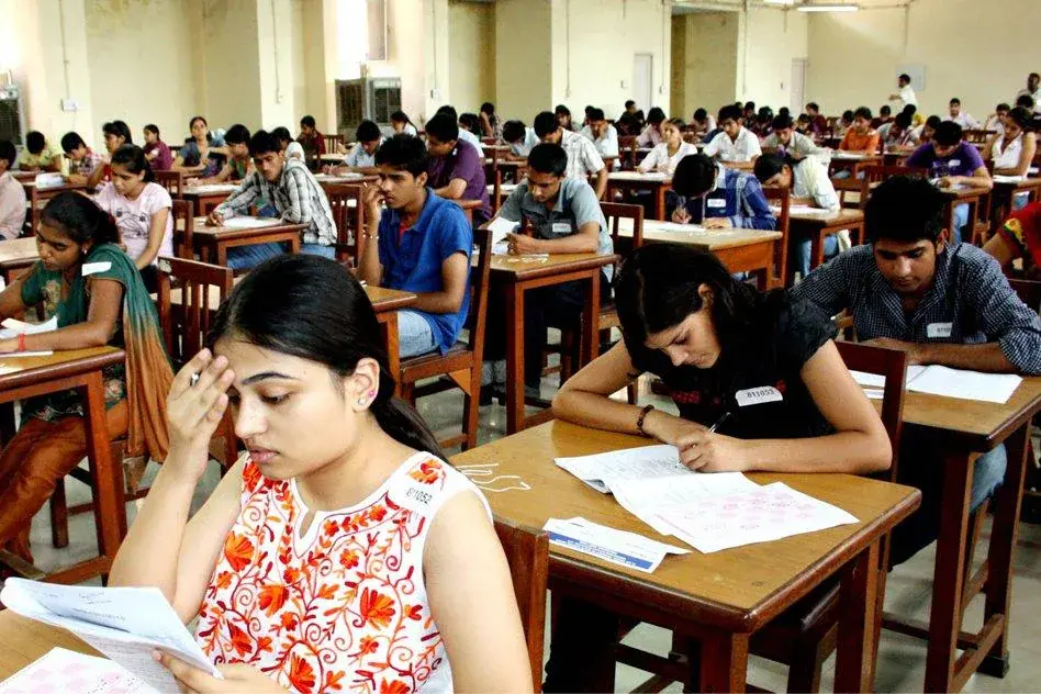 A large exam hall filled with students seated at wooden desks in rows, writing on answer sheets, with a woman in the foreground pausing to think.