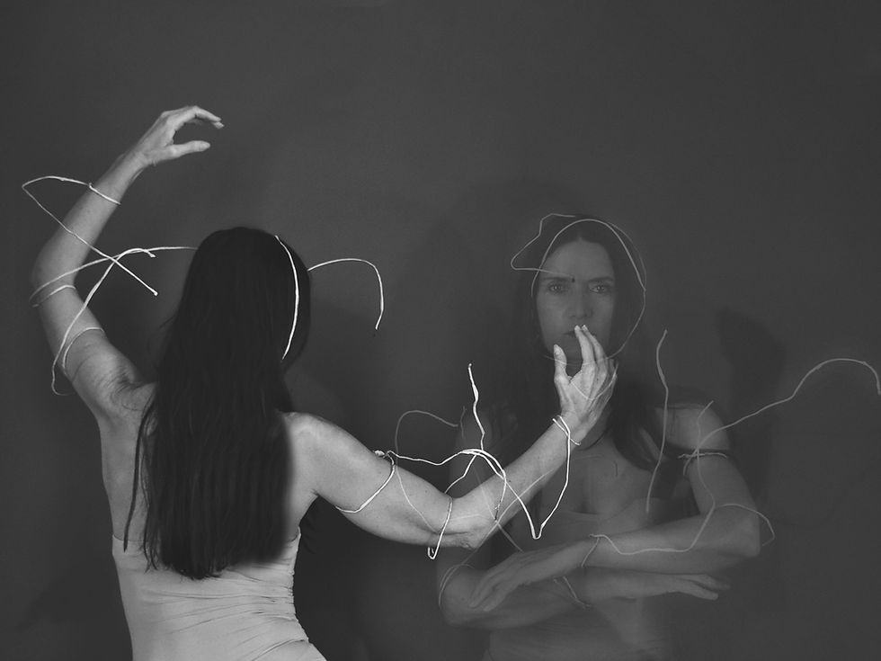 Fotografia em preto e branco de uma mulher de costas, parcialmente envolta por fios brancos que serpenteiam pelo seu corpo e se estendem pelo espaço. À frente dela, em sobreposição ou reflexo, vê-se o rosto da mesma mulher encarando a câmera com expressão séria, uma mão parcialmente cobrindo seus lábios. A cena sugere um jogo entre presença e projeção, com elementos visuais que evocam tensão, introspecção e dualidade entre gesto e silêncio. A composição é etérea, marcada por linhas sinuosas e uma atmosfera densa.