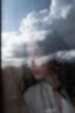 Fotografia artística retrata uma mulher de cabelos escuros e expressão contemplativa, vista através de um vidro coberto por gotas de chuva. O reflexo de nuvens no céu se sobrepõe ao seu rosto, criando uma fusão poética entre o ambiente externo e sua presença. Vestindo uma blusa clara, ela olha para o lado, com o olhar perdido, evocando sentimentos de introspecção e melancolia. A composição explora camadas de transparência, reflexo e textura, unindo o real e o efêmero em uma atmosfera delicada e emocionalmente densa, ideal para contextos de fotografia contemporânea e narrativa visual.