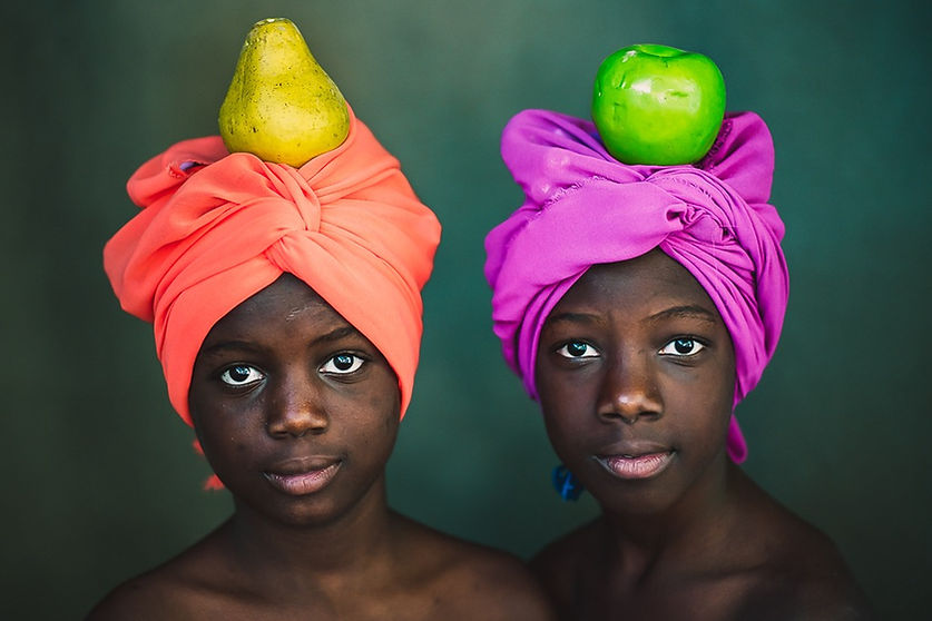 Portrait of two Black girls with colorful headwraps, one in orange with a pear on top and the other in pink with a green apple, against a dark background.