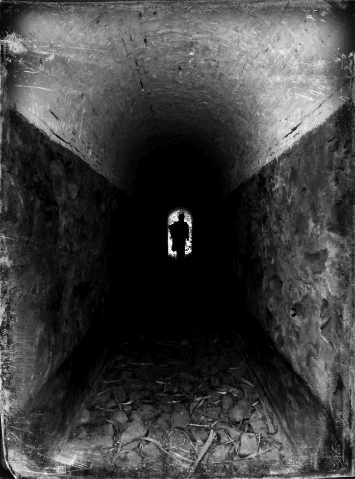 Black and white photograph of a narrow, dark tunnel with stone walls and a curved ceiling. At the far end, in the small illuminated opening, the silhouette of a standing figure is visible, sharply contrasted against the light