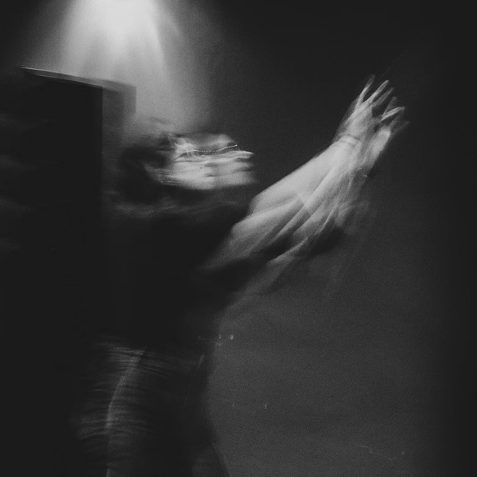 Imagem em preto e branco de uma figura humana em movimento, capturada com longa exposição, o que resulta em um rastro fantasmagórico e múltiplas sobreposições do corpo e dos braços estendidos para o alto. A cena transmite um sentido de urgência, transcendência ou busca, como se a figura estivesse tentando alcançar algo invisível. A luz suave no canto superior ilumina parcialmente o rosto desfocado, reforçando a atmosfera etérea e enigmática da composição. A imagem sugere um estado liminar entre presença e ausência, evocando sensações de desmaterialização, instabilidade e transcendência do gesto.