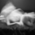 Fotografia em preto e branco de longa exposição mostra um corpo nu deitado em movimento, criando uma figura difusa e espectral. As linhas borradas sugerem fluxo e instabilidade, como se a imagem oscilasse entre presença e ausência. O fundo escuro acentua o contraste e reforça a atmosfera onírica e fantasmagórica.