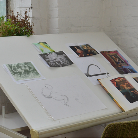 Mesa de trabalho com imagens, desenhos e livro aberto, representando processo de seleção e construção de portfólio artístico.