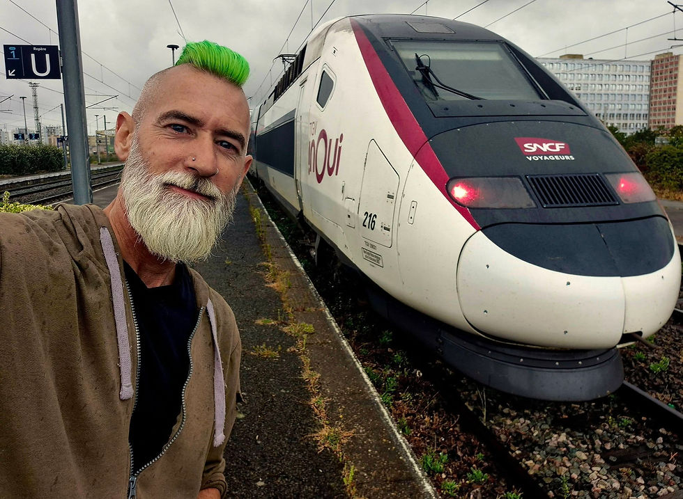 Selfie of a man with neon green hair and white beard beside an SNCF TGV InOui train at a rainy Boulogne sur Mer railway station.
