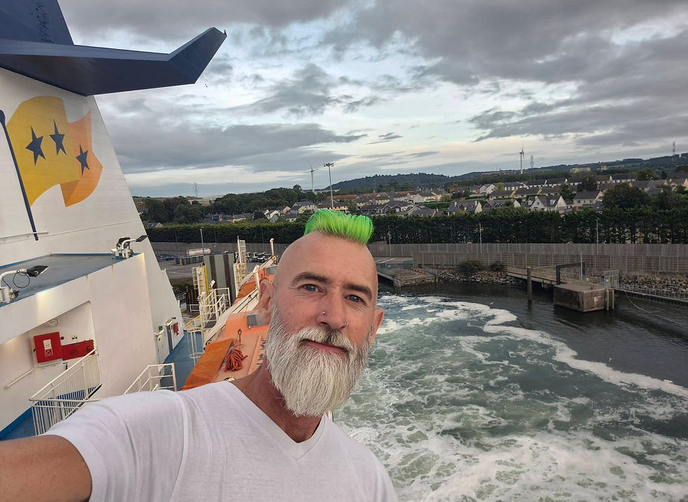 Departing Ringaskiddy Cork on Hibernia Line ferry. Man with neon green mohawk takes a selfie on a ferry deck above a churning harbour, with cloudy sky and coastal homes behind.
