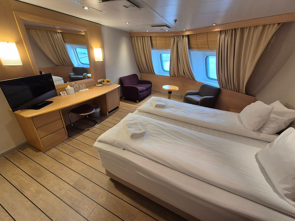 Cosy Hibernia Line deluxe cabin with twin beds, desk, TV, chairs, and curtained porthole windows in warm wood tones