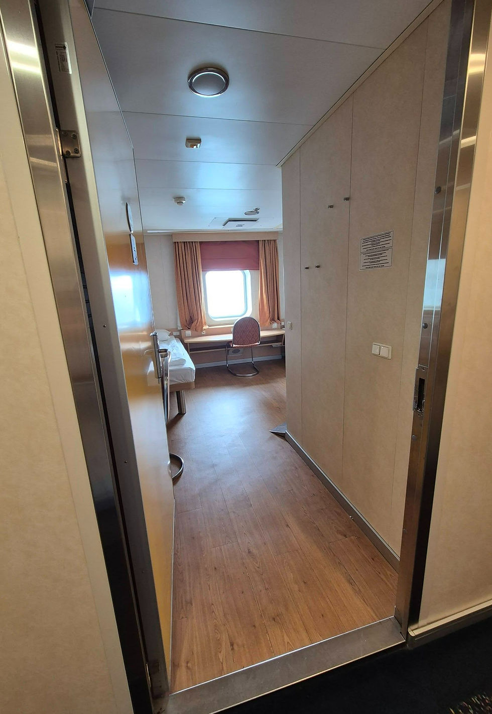Wheelchair accessible cabin on Hibernia Line ferry with wood floor, bed, desk, chair, and bright window framed by pink curtains