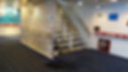 Staircase in a ship's interior with white railings and blue steps. Welcome sign on wall. Carpeted floor. Bright, clean atmosphere.