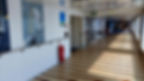 Sunlit ship corridor with striped floor and sailing ship mural. Two people walk away. Blue signs and red fire extinguisher on white walls.