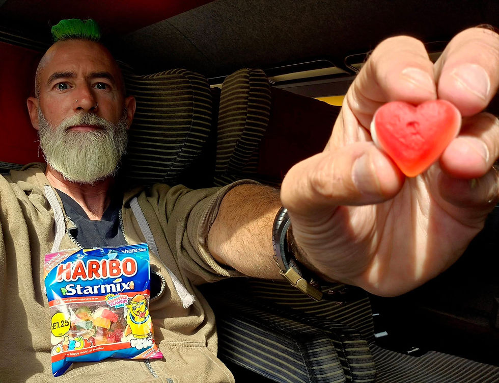 Bearded man with green mohawk in an SNCF TGV highspeed train holds up a red heart candy; Haribo Starmix bag on his chest.