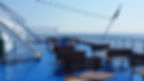 Empty ferry deck with wooden benches and blue flooring, under a clear sky with ocean view. A flag waves gently in the breeze.