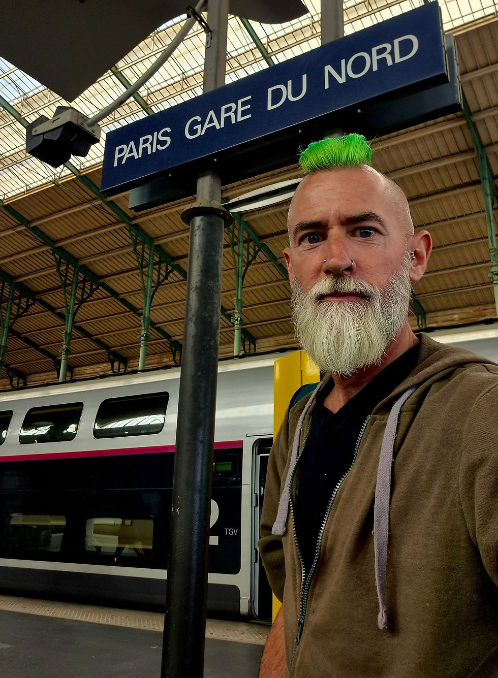 Man with neon green mohawk and white beard stands at Paris Gare du Nord beside a TGV train, looking serious.