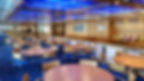 Empty dining area with wooden tables, blue chairs, and a blue-lit ceiling. Walls have posters. High chairs are lined up on the right.