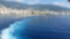 Blue sea with foamy trail from a ferry, coastal city of Bastia in the background with colourful buildings and mountain landscape under a clear sky.