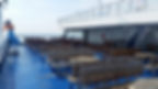 Empty ferry deck with wooden benches and tables on a blue floor. A large penguin statue wearing a sailor hat stands at the railing.