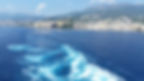 View of a coastal city on Corsica with mountains in the background. Blue ocean with white waves create patterns in the foreground. Clear, sunny day.