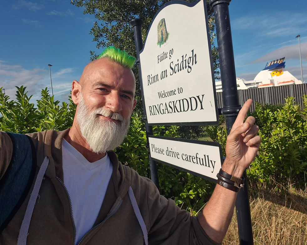 Man with green mohawk points at Ringaskiddy welcome sign reading Faílte go Rinn an Scidigh and Please drive carefully. Hibernia Line ferry in the background
