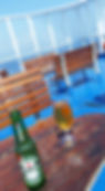 Beer bottle and glass on a wooden table on a sunny ferry deck. Blue floor, wooden benches, and clear sky visible. Relaxing ambiance.