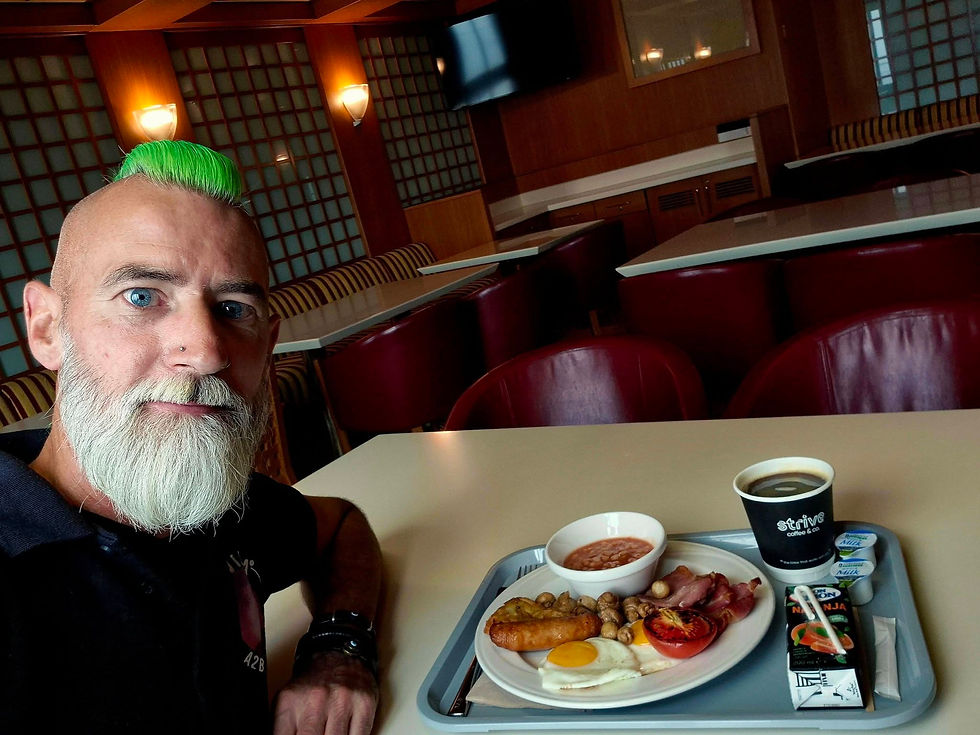Bearded man with bright green mohawk takes a breakfast selfie at a dinner table aboard a Hibernia Line ferry with breakfast tray and coffee in an empty warm-toned room