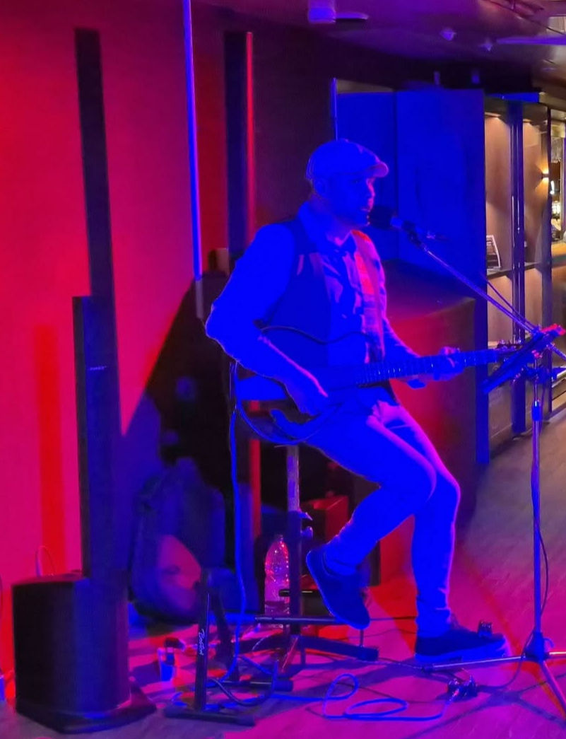 Man in a cap plays guitar and sings into a mic on a small stage on DFDS King Seaways ferry, bathed in blue and pink lights at a bar.