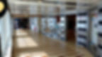 Hallway on a ship with striped floors. A man and girl walk towards a shop. Glass doors, light reflections, and posters in the background.