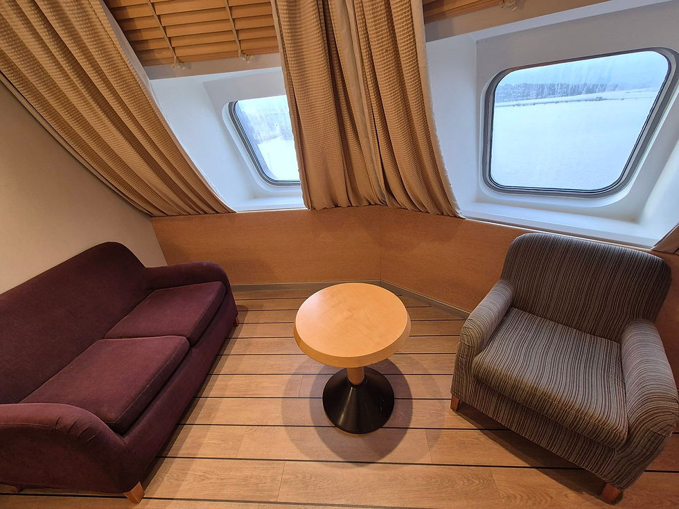 Hibernia Line deluxe cabin lounge with a plum sofa, striped armchair, and round table by large windows overlooking St Patrick bow