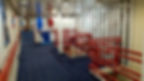 Hallway with blue carpet, red railings, white walls, and doors. Signs say “PURDY CORRDOR” and “NO ENTRY.” Bright overhead lights.