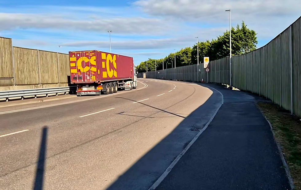 Semi-truck hauling a red and yellow container around a curved road in Ringaskiddy Cork beside sound barriers under a blue, cloudy sky.