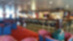 Spacious bar area with red carpet, blue and red chairs, a man near the bar, and warm lighting. Bottles visible on shelves behind the bar.