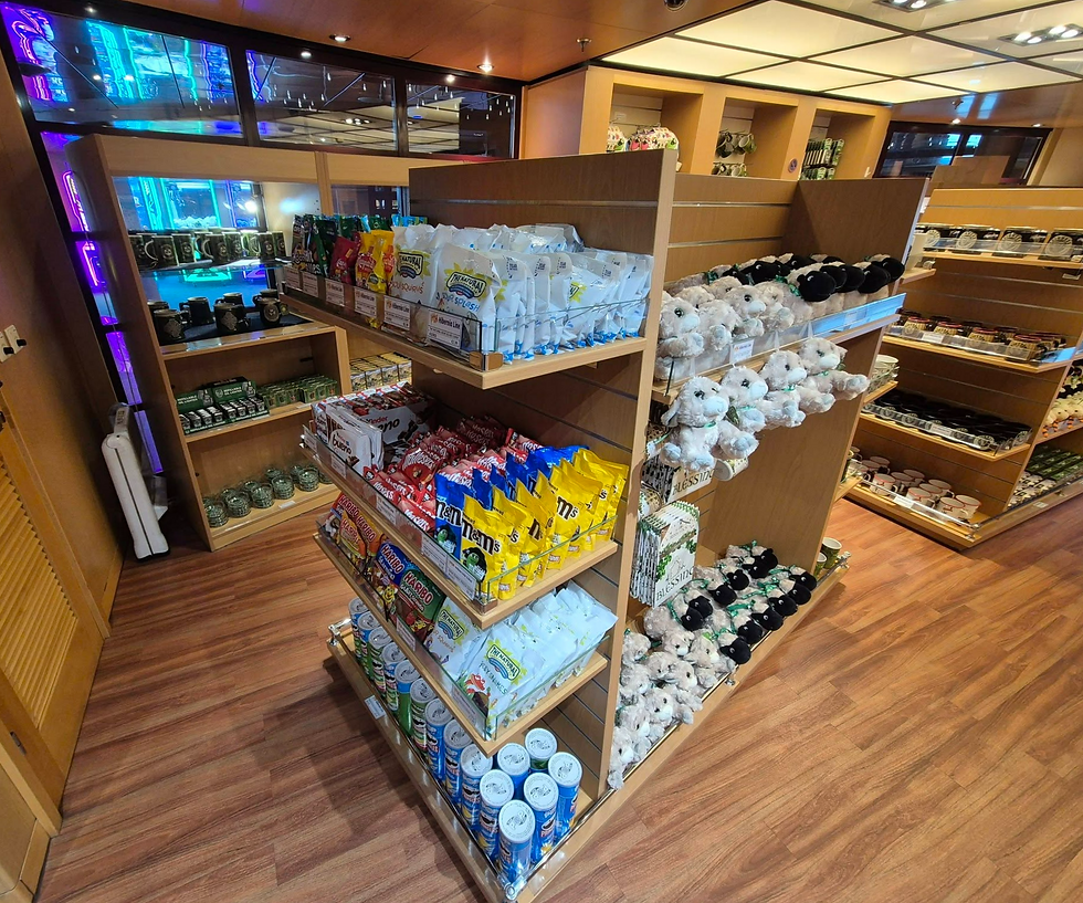 Gift shop display with colourful snacks, mugs, and plush teddy bears on wooden shelves in a bright wood-floored store aboard a Hibernia Line ferry