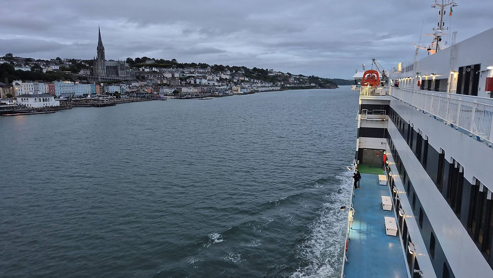 Hibernia Line ferry sailing past Cobh in Ireland with hillside houses and a tall church spire at dusk, wake trailing behind.