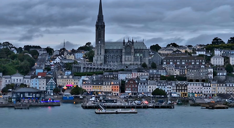 Dusk view of Cobh waterfront town with colorful houses, St Colman's cathedral spire, and a small ferry on calm water under cloudy skies.