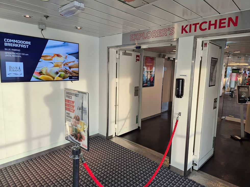 DFDS ferry breakfast entrance labeled EXPLORER'S KITCHEN, with menu screens, open doors, and red rope barrier in bright hallway.