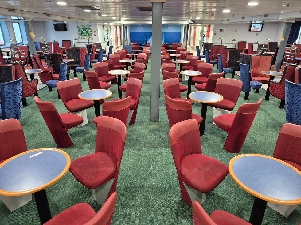 Empty ferry seating area with rows of red and blue chairs and circular tables from the former HSS Stena Discovery, set on green carpet. Brightly lit, with windows and signage.