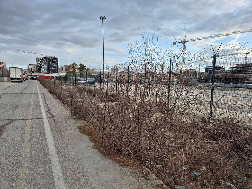 Urban scene with trucks parked on a road. Barbed wire fence and barren bushes in the foreground. Cloudy sky, cranes, and buildings in the background.