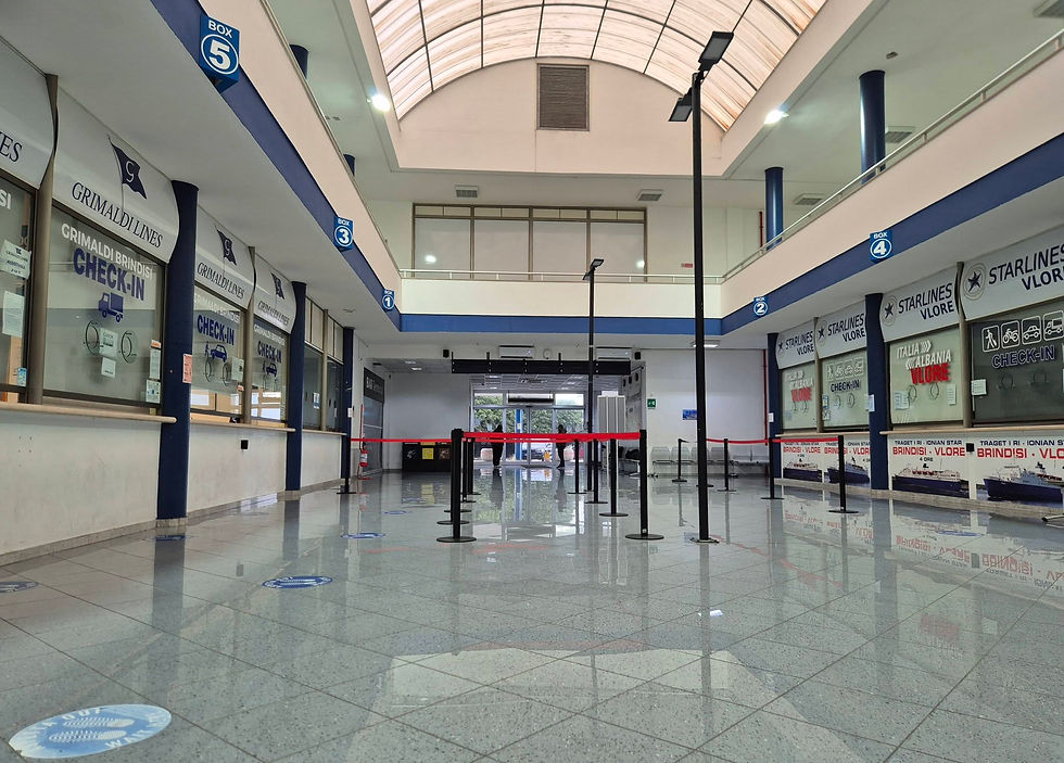 Empty ferry terminal hall with check-in desks for Grimaldi Lines and Starlines Vlore. Red barriers, blue signs, and a skylight ceiling.