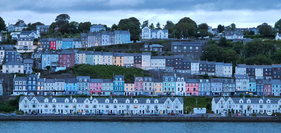 Colorful terraced houses on Cobh hillside beside the water at dusk, under a cloudy sky, with lights glowing in windows.